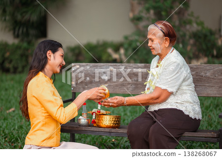 Young woman kneeling to pour water into an elderly woman hands during a traditional Thai Songkran ceremony on a bench outdoors, showing humility, respect for elders and meaningful family care. Young woman kneeling to pour water into an elderly woman hands during a traditional Thai Songkran ceremony on a bench outdoors, showing humility, respect for elders and meaningful family care. 138268067
