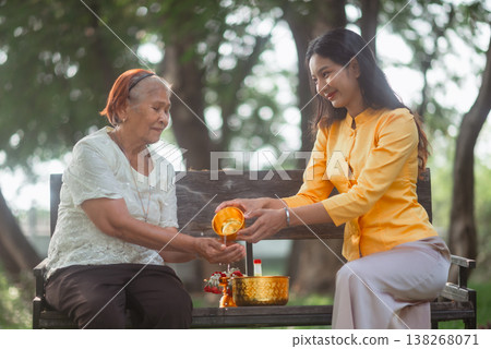 Young woman pouring water into an elderly woman hands on a wooden bench during Songkran, showing Thai family values, respect for elders and a peaceful traditional New Year blessing ritual. 138268071