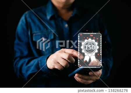 A person using a smartphone displays a glowing holographic ISO certification badge symbolizing quality assurance, compliance, and international standards in modern digital business management. 138268276