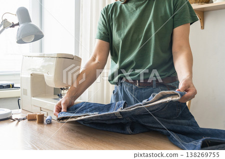 Woman standing at a desk, smoothing out a pair of jeans, preparing for sewing or alteration with a sewing machine nearby Woman standing at a desk, smoothing out a pair of jeans, preparing for sewing or alteration with a sewing machine nearby 138269755