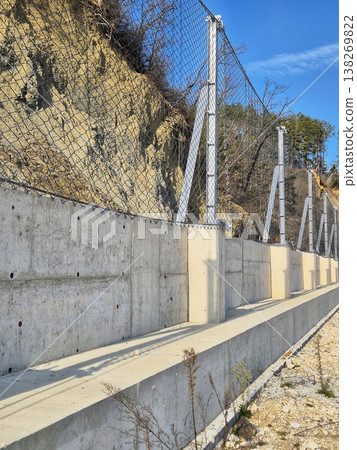 Concrete retaining wall with steel mesh fence for landslide and rockfall protection on a steep slope. 138269822