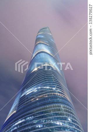 A view looking up at a spiral glass skyscraper that twists and rises towards a pink, cloudy sky. A view looking up at a spiral glass skyscraper that twists and rises towards a pink, cloudy sky. 138270027