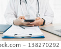 Doctor in white coat uses smartphone at clinic desk with stethoscope, clipboard and laptop, representing telemedicine communication, digital records and health insurance claim support. 138270292