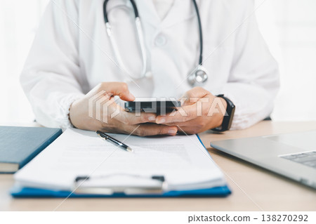 Doctor in white coat uses smartphone at clinic desk with stethoscope, clipboard and laptop, representing telemedicine communication, digital records and health insurance claim support. 138270292