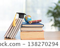 Stack of textbooks with graduation cap and red tassel beside pencil holder and globe on desk in bright modern room. Education, study, elearning, academic success, back to school concept. 138270294