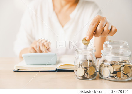 Hand dropping coin into glass savings jar on desk with scattered coins, notebook and phone. Budget planning, personal finance, frugal living, investment goals, future security. investment goals Hand dropping coin into glass savings jar on desk with scattered coins, notebook and phone. Budget planning, personal finance, frugal living, investment goals, future security. investment goals 138270305