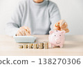 Hand placing a coin into a pink piggy bank beside stacks of money while writing budget notes at home. Personal finance, savings plan, budgeting, investment, long-term future security goals. 138270306