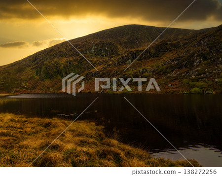 Lake shore with dry golden grass and autumn heather covered slopes reflecting in dark water. Lake shore with dry golden grass and autumn heather covered slopes reflecting in dark water. 138272256