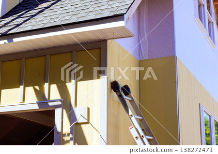 Ladder is set against building as workers carry out construction tasks installing primed smooth fiber siding Ladder is set against building as workers carry out construction tasks installing primed smooth fiber siding 138272471