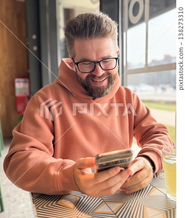 Smiling man using smartphone while sitting in a cafe near a large window with a drink on the table. Leisure lifestyle, digital communication, remote work culture, relaxation and modern urban social Smiling man using smartphone while sitting in a cafe near a large window with a drink on the table. Leisure lifestyle, digital communication, remote work culture, relaxation and modern urban social 138273310