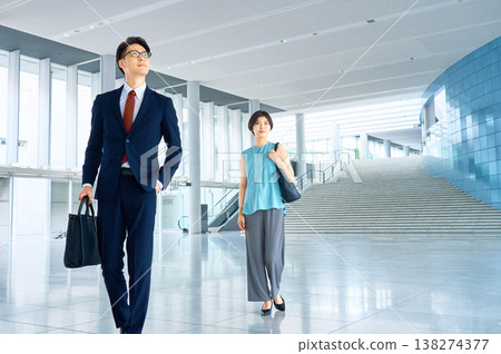 A businessman and businesswoman stride confidently through a modern office building. A businessman and businesswoman stride confidently through a modern office building. 138274377