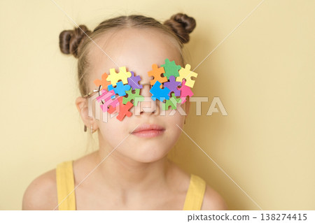 A child wearing glasses made from a variety of pieces of bright multicolored puzzles, demonstrating support and awareness of autism. A girl on a yellow studio background 138274415