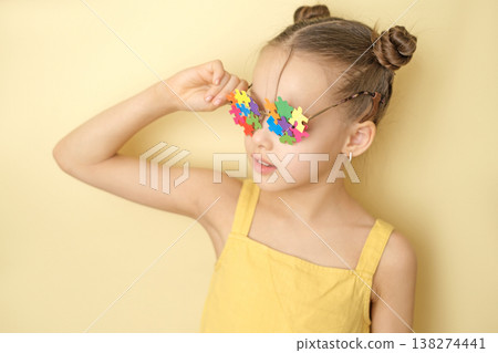 A cute girl with a funny hairstyle wears glasses decorated with multicolored puzzle pieces as a sign of support for people with autism, education and social awareness. 138274441