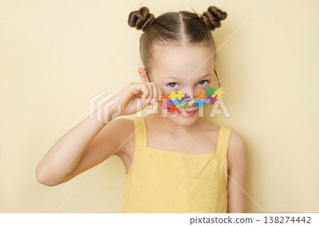 The cute girl lowered glasses decorated with pieces of colorful puzzles, looking suspiciously and smiling at the camera. World Autism Awareness Day. Education and social equality 138274442