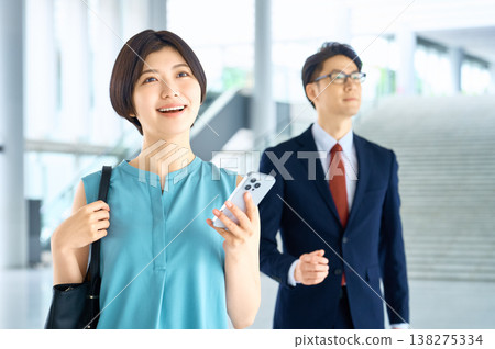 一位女商人微笑著站在辦公大樓裡,手裡拿著智慧型手機,背景中一位穿著西裝的男士正在拍照。 一位女商人微笑著站在辦公大樓裡,手裡拿著智慧型手機,背景中一位穿著西裝的男士正在拍照。 138275334