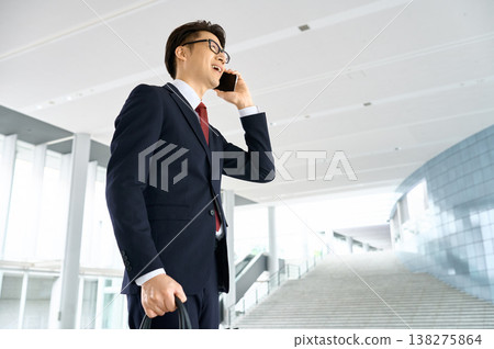 A Japanese businessman smiles and uses his smartphone to make a call in the lobby of an office building. 138275864