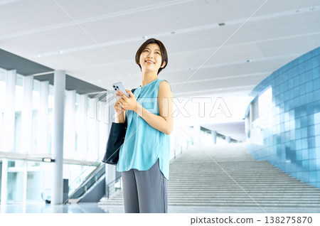 一位女商人面帶微笑地站在辦公大樓大廳裡，手裡拿著她的智慧型手機。 138275870