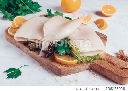 Fresh squid prepared for cooking, surrounded by herbs and lemon, placed on a wooden chopping board in a bright kitchen setting Fresh squid prepared for cooking, surrounded by herbs and lemon, placed on a wooden chopping board in a bright kitchen setting 138278938