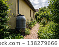 Rainwater barrel collects water next to a rural house with yellow wall 138279666
