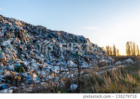 A towering mountain of garbage spreads across the landfill, reflecting the fading light of sunset while nature stands in contrast nearby 138279718