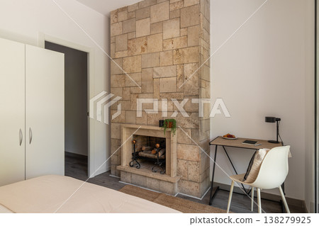 A modern bedroom featuring a stone fireplace, white wardrobe, and a desk with a chair. The room has neutral colors and a minimalist design. A modern bedroom featuring a stone fireplace, white wardrobe, and a desk with a chair. The room has neutral colors and a minimalist design. 138279925