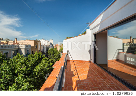 A modern balcony view in Barcelona. The scene includes a clear blue sky, terracotta tiles, and green trees lining the street. Urban architecture is visible in the background. A modern balcony view in Barcelona. The scene includes a clear blue sky, terracotta tiles, and green trees lining the street. Urban architecture is visible in the background. 138279926
