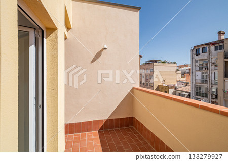 A sunny balcony with a terracotta floor and beige walls. The view includes nearby buildings under a clear blue sky. 138279927