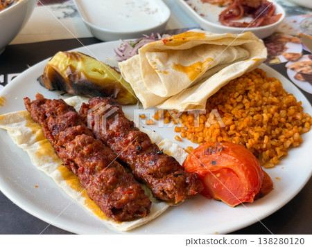 Adana kebab served with bulgur pilaf, lavash, onion salad and roasted vegetables on plate. Traditional Turkish restaurant meal, hearty lunch portion and menu ready savory presentation Adana kebab served with bulgur pilaf, lavash, onion salad and roasted vegetables on plate. Traditional Turkish restaurant meal, hearty lunch portion and menu ready savory presentation 138280120