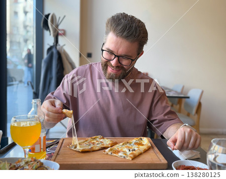 Smiling man lifting a slice of cheese turkish pide with melted topping in a bright cafe. Casual food enjoyment, authentic dining experience, weekend lunch mood and restaurant lifestyle scene 138280125