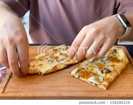 Hands reaching for cheese and mushroom pide slices on a wooden serving board. Casual meal interaction, shared lunch concept and appetizing restaurant food moment with human presence 138280129