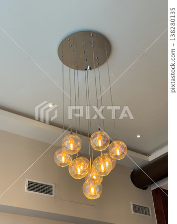 Modern glass pendant chandelier with glowing bulbs suspended from a circular ceiling mount. Contemporary interior lighting design, warm hospitality ambience and decorative architectural detail 138280135