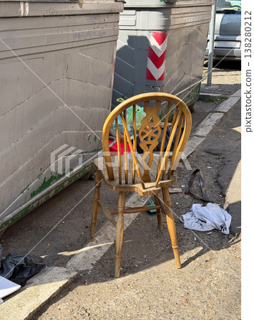 Abandoned wooden chair standing beside dumpsters and scattered litter in harsh sunlight. Urban neglect, illegal dumping, public sanitation issue and social documentary atmosphere in a backstreet 138280212