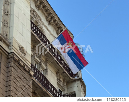 Serbian national flag mounted on an ornate historic building facade against a clear blue sky. State identity, civic symbolism, official representation and architectural heritage in an urban capital Serbian national flag mounted on an ornate historic building facade against a clear blue sky. State identity, civic symbolism, official representation and architectural heritage in an urban capital 138280213
