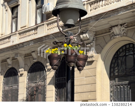 Historic street lamp with flower planters against an ornate city facade in bright daylight. Urban streetscape decoration, architectural heritage detail and civic beautification in a downtown setting 138280214