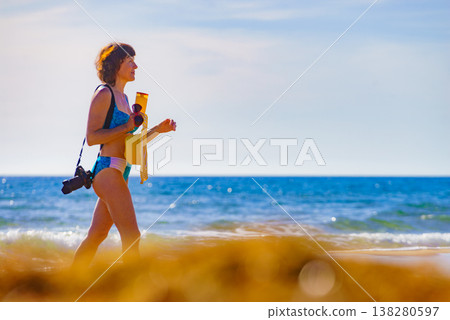 Woman in bikini with camera walk on beach Woman in bikini with camera walk on beach 138280597