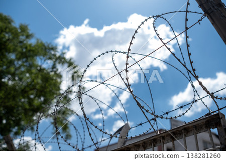 Barbed Wire Stretches Against a Bright Blue Sky With Clouds in a Secure Location Indicating Barriers and Confinement Barbed Wire Stretches Against a Bright Blue Sky With Clouds in a Secure Location Indicating Barriers and Confinement 138281260
