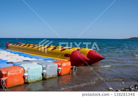 Bright Yellow Banana Boat Resting at the Shoreline of a Clear Blue Ocean on a Sunny Day, Ready for Fun Water Sports Bright Yellow Banana Boat Resting at the Shoreline of a Clear Blue Ocean on a Sunny Day, Ready for Fun Water Sports 138281348