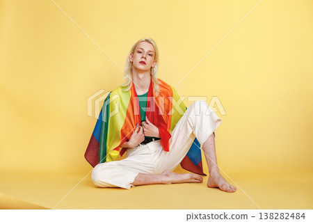 Young blonde man with make up on his face and rainbow flag posing for a photo. Man wearing fashionable green top and has red lips. Man isolated on yellow background. Young blonde man with make up on his face and rainbow flag posing for a photo. Man wearing fashionable green top and has red lips. Man isolated on yellow background. 138282484