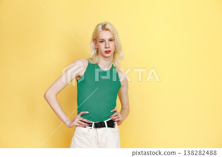 Young blonde man with make up on his face posing for a photo. Man wearing fashionable green top and has red lips. Man isolated on yellow background. Young blonde man with make up on his face posing for a photo. Man wearing fashionable green top and has red lips. Man isolated on yellow background. 138282488