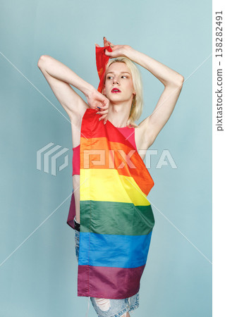 Young blonde man with make up on his face and rainbow flag posing for a photo. Man wearing fashionable pink t-shirt and has red lips. Man isolated on blue background. Young blonde man with make up on his face and rainbow flag posing for a photo. Man wearing fashionable pink t-shirt and has red lips. Man isolated on blue background. 138282491