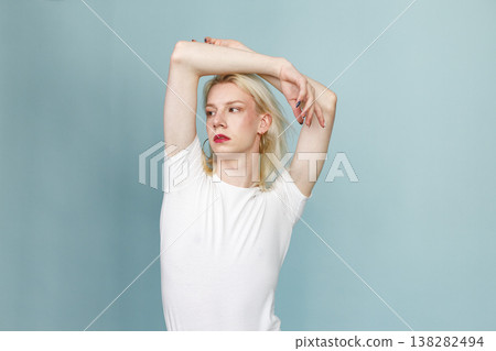 Young blonde man with make up on his face posing for a photo. Man wearing fashionable white t-shirt and has red lips. Man isolated on blue background. Young blonde man with make up on his face posing for a photo. Man wearing fashionable white t-shirt and has red lips. Man isolated on blue background. 138282494