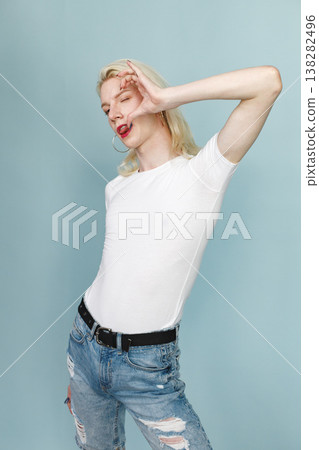 Young blonde man with make up on his face posing for a photo. Man wearing fashionable white t-shirt and has red lips. Man isolated on blue background. Young blonde man with make up on his face posing for a photo. Man wearing fashionable white t-shirt and has red lips. Man isolated on blue background. 138282496