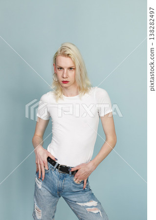 Young blonde man with make up on his face posing for a photo. Man wearing fashionable white t-shirt and has red lips. Man isolated on blue background. Young blonde man with make up on his face posing for a photo. Man wearing fashionable white t-shirt and has red lips. Man isolated on blue background. 138282497