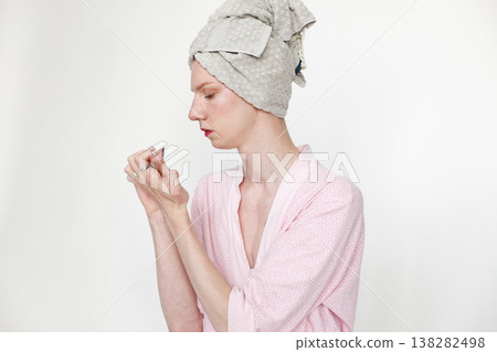 Young man with make up on his face doing manicure. Man wearing pink dressing gown and has a towel on his head. Man isolated on white background. Young man with make up on his face doing manicure. Man wearing pink dressing gown and has a towel on his head. Man isolated on white background. 138282498