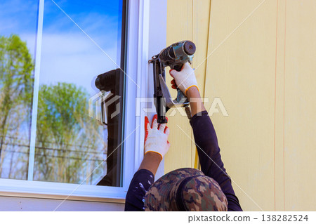 Carpenter is fitting window frame using nail gun on building while working outside Carpenter is fitting window frame using nail gun on building while working outside 138282524
