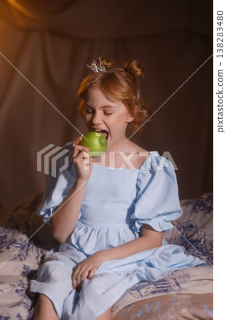 Little girl in blue dress with crown eating green apple, princess concept, studio drapery background Little girl in blue dress with crown eating green apple, princess concept, studio drapery background 138283480