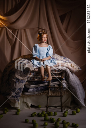 Little girl in blue dress sitting on stacked mattresses with apples, princess concept, studio drapery Little girl in blue dress sitting on stacked mattresses with apples, princess concept, studio drapery 138283481