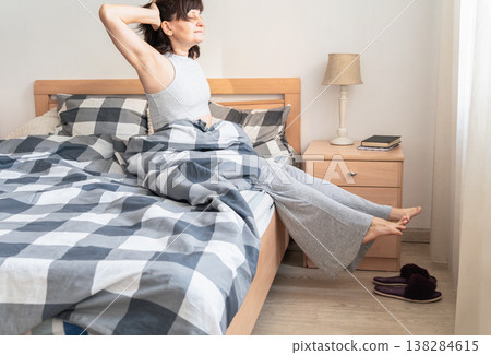An adult woman waking up in the morning, sitting on bed and stretching arms, feeling refreshed and ready for the day ahead. Authentic lifestyle 138284615
