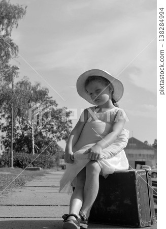 Young girl in a hat sits on an old suitcase, waiting patiently outdoors Young girl in a hat sits on an old suitcase, waiting patiently outdoors 138284994