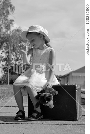 Young girl in a hat and dress sits on a suitcase with a stuffed toy, waiting for a journey Young girl in a hat and dress sits on a suitcase with a stuffed toy, waiting for a journey 138285000